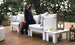 A woman enjoys ultimate relaxation on a wooden deck, wrapped in a dark shawl and smiling serenely amidst the trees. She sits on the Ledge Lounger Mainstay Sectional Relaxed Endcap, where candles glow gently in lanterns beside a small stack of books on an elegant table.