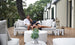 A couple relaxes on a Mainstay Sectional Relaxed Right Corner by Ledge Lounger, surrounded by lush greenery. They smile, leaning against each other amidst white and gray cushions. A round coffee table and decorative lanterns complete this enchanting scene.