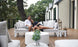 A couple relaxes on a Mainstay Sectional Relaxed Right Corner by Ledge Lounger, surrounded by lush greenery. They smile, leaning against each other amidst white and gray cushions. A round coffee table and decorative lanterns complete this enchanting scene.
