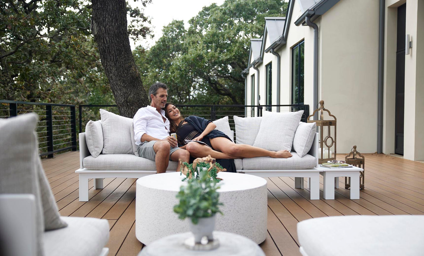 A couple relaxes on a modern deck, enjoying the comfort of the Mainstay Sectional Relaxed Middle by Ledge Lounger. Theyre lounging on a white sofa amid lush greenery, with a round white table and potted plant adding charm, while a large house with stylish railing looms in the background.