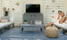 A couple sits on an outdoor sofa enjoying drinks in a cozy patio setup, focused on all-weather durability. The scene includes a TV on a stand, the Mainstay Rectangular Coffee Table with a plant, a large ice bucket, and woven pouf. White slatted walls and geometric rug add modern flair.