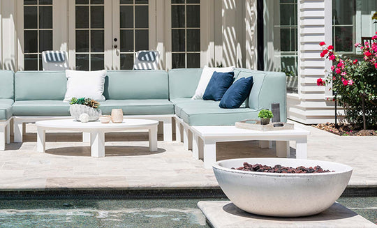 An outdoor patio showcases a light green Ledge Lounger sectional sofa paired with a Mainstay Sectional Left Armchair and white cushions, complemented by a white coffee table, decor, and round fire pit. Glass doors in the background and pink flowers to the right enhance the scene.