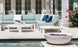 An outdoor patio showcases a light green Ledge Lounger sectional sofa paired with a Mainstay Sectional Left Armchair and white cushions, complemented by a white coffee table, decor, and round fire pit. Glass doors in the background and pink flowers to the right enhance the scene.