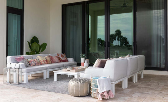 The patio showcases a Ledge Lounger Mainstay Sectional with modern white sofas, vibrant patterned cushions, and a low white table. A potted plant, metallic pouf, and wicker basket enhance the decor. Large glass doors and a textured rug complete the cozy ambiance.