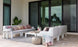 The patio showcases a Ledge Lounger Mainstay Sectional with modern white sofas, vibrant patterned cushions, and a low white table. A potted plant, metallic pouf, and wicker basket enhance the decor. Large glass doors and a textured rug complete the cozy ambiance.