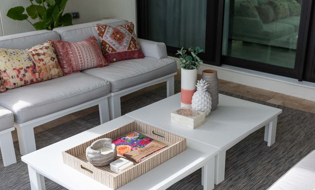 A modern outdoor seating area includes a Ledge Lounger Mainstay Sectional Right Armchair with white sofas, colorful patterned pillows, and a white coffee table. Potted plants decorate the gray rug while a wicker tray with magazines, a candle, and decor adds charm.