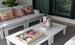 A cozy outdoor patio showcases a Mainstay Sectional Left Armchair by Ledge Lounger, featuring white seating and colorful patterned cushions. A large white coffee table displays a wicker tray with books, a candle, and decorative items like a pineapple ornament and plant-filled vases.