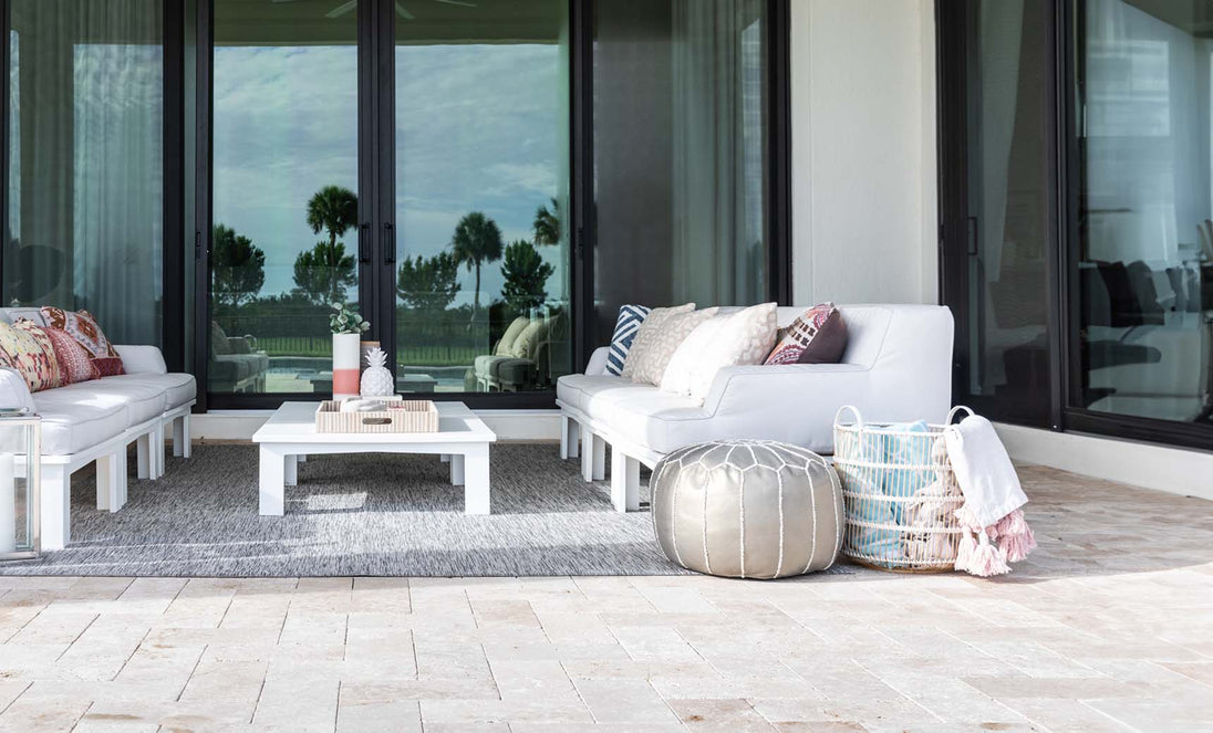 The outdoor patio includes Ledge Lounger white cushioned seating with a Mainstay Sectional Right Armchair, centered around a sleek white coffee table on a gray rug. Two poufs and a basket with blankets sit on the stone-tiled floor. Glass doors reflect palm trees under a cloudy sky.