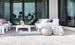 The outdoor patio includes Ledge Lounger white cushioned seating with a Mainstay Sectional Right Armchair, centered around a sleek white coffee table on a gray rug. Two poufs and a basket with blankets sit on the stone-tiled floor. Glass doors reflect palm trees under a cloudy sky.