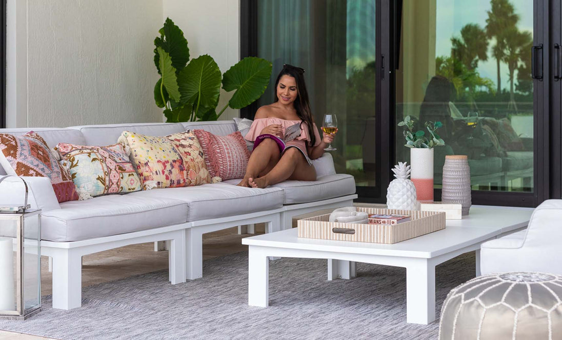 A woman lounges in a Mainstay Sectional Right Armchair by Ledge Lounger, accessorized with a vibrant cushion set, sipping her drink while reading a magazine. A white coffee table holds decorative items nearby, surrounded by lush green plants and sleek glass doors in the background.