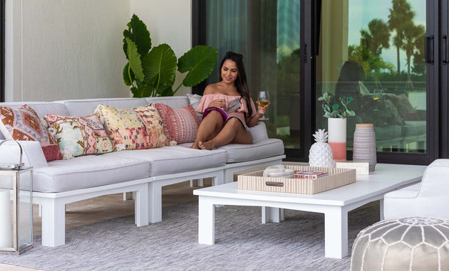 A woman lounges in a Mainstay Sectional Right Armchair by Ledge Lounger, accessorized with a vibrant cushion set, sipping her drink while reading a magazine. A white coffee table holds decorative items nearby, surrounded by lush green plants and sleek glass doors in the background.