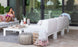 A woman relaxes on the Mainstay Sectional Right Armchair by Ledge Lounger, sipping her drink and perusing a magazine. The white outdoor furniture, with its stylish cushion set and pouf, complements the serene pool view and lush greenery through the glass doors.