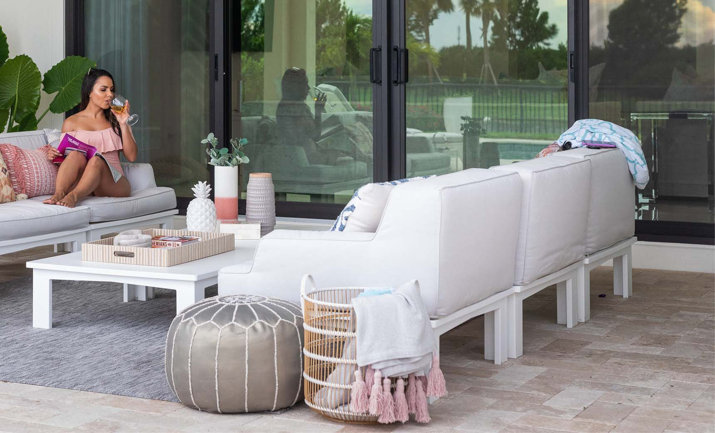 A woman in a pink dress sits on a Ledge Lounger Mainstay Sectional Left Armchair, sipping a drink and holding a book. The patio has white sofas with cushions, a pouf, and a basket of blankets. A coffee table holds a tray, while large glass doors mirror the lush greenery outside.