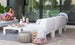 A woman in a pink dress sits on a Ledge Lounger Mainstay Sectional Left Armchair, sipping a drink and holding a book. The patio has white sofas with cushions, a pouf, and a basket of blankets. A coffee table holds a tray, while large glass doors mirror the lush greenery outside.