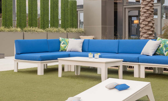 Outdoor seating showcases a Ledge Lounger Mainstay Sectional Corner with blue cushions and white tables on grassy turf. Accentuated by decorative plants and a palm tree, the space is completed with soft green and patterned pillows for limitless arrangement options.