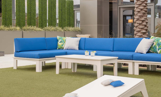 Outdoor seating showcases a Ledge Lounger Mainstay Sectional Corner with blue cushions and white tables on grassy turf. Accentuated by decorative plants and a palm tree, the space is completed with soft green and patterned pillows for limitless arrangement options.