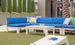 Outdoor seating showcases a Ledge Lounger Mainstay Sectional Corner with blue cushions and white tables on grassy turf. Accentuated by decorative plants and a palm tree, the space is completed with soft green and patterned pillows for limitless arrangement options.