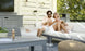 A couple smiles on an outdoor couch, both in sunglasses. The woman holds a glass with an ice bucket on the table. Nearby is a Ledge Lounger Mainstay Sectional Corner with three-piece cushions, a TV, and potted plant set against lush greenery creates a bright and relaxed scene.