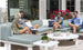 Four people sit outside on a Ledge Lounger Mainstay Sectional Corner, chatting casually atop its three-piece cushions. The area is adorned with potted plants and flowers, while a building with large windows in the background reflects sunlight onto them.