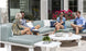 Four people sit outside on a Ledge Lounger Mainstay Sectional Corner, chatting casually atop its three-piece cushions. The area is adorned with potted plants and flowers, while a building with large windows in the background reflects sunlight onto them.
