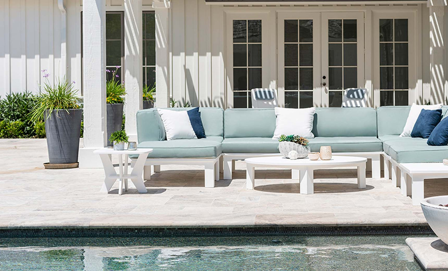 In a patio setting by the pool, the Mainstay Sectional Corner by Ledge Lounger is arranged in white and blue hues around white coffee tables. Tall planters add greenery, while a white building with large windows sets the backdrop, offering limitless arrangement options.
