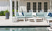 In a patio setting by the pool, the Mainstay Sectional Corner by Ledge Lounger is arranged in white and blue hues around white coffee tables. Tall planters add greenery, while a white building with large windows sets the backdrop, offering limitless arrangement options.
