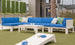 The outdoor seating area boasts a Ledge Lounger Mainstay Sectional Middle with a large blue L-shaped sofa adorned with tropical and white pillows. A white coffee table and cornhole board sit on the green lawn, complemented by tall planters and a palm tree in the serene backdrop.