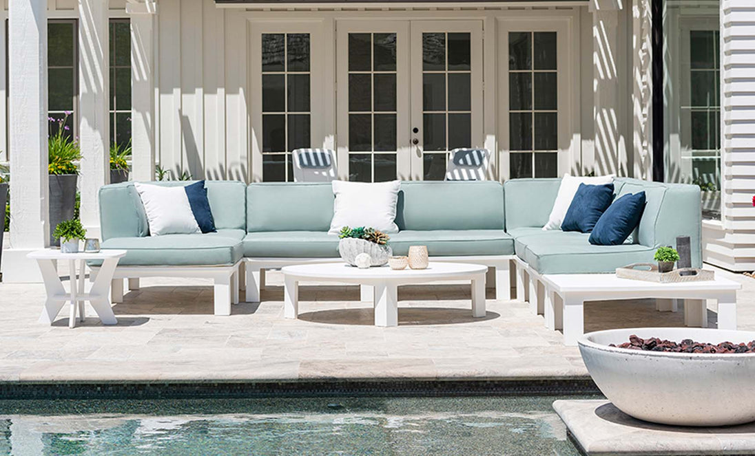 An outdoor patio boasts a Ledge Lounger Mainstay Sectional Middle in light grey with white and navy cushions. White side and coffee tables with decor enhance the scene, complemented by French doors, potted plants, a pool, and a fire bowl completing the ambience.