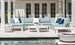 An outdoor patio boasts a Ledge Lounger Mainstay Sectional Middle in light grey with white and navy cushions. White side and coffee tables with decor enhance the scene, complemented by French doors, potted plants, a pool, and a fire bowl completing the ambience.