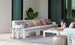 A stylish patio showcases the Mainstay Sectional Middle by Ledge Lounger, featuring a white-cushioned sofa with colorful patterned pillows. A white coffee table displays decor items, flanked by two lanterns and a large green plant on neutral-toned tiles near glass doors.