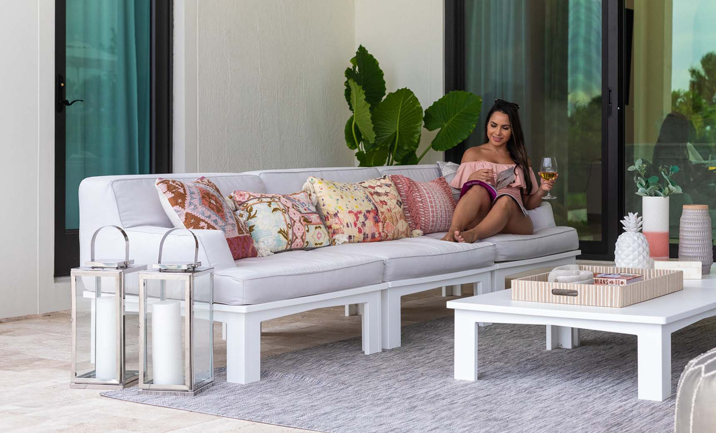 A woman unwinds on a Mainstay Sectional Middle by Ledge Lounger, its pre-assembled base featuring a vibrant cushion set. She sips her drink and is engrossed in her book or tablet, surrounded by large plants, glass candle lanterns, and patio decor creating a serene ambiance.