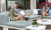 In a sunny garden, two women and a man relax on light blue Mainstay Sectional Ottomans by Ledge Lounger. They smile amid potted plants and vivid pink flowers, enjoying the sectionals versatile arrangements for perfect relaxation.