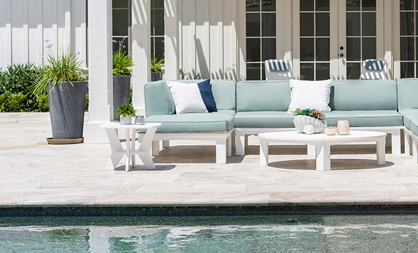Mainstay light blue and white sectional sofa by Ledge Lounger adorns the poolside patio. A round white table with decor complements the ambiance, inviting endless configurations. Large potted plants encircle, while glass doors mirror the serene pool ahead.