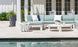 Mainstay light blue and white sectional sofa by Ledge Lounger adorns the poolside patio. A round white table with decor complements the ambiance, inviting endless configurations. Large potted plants encircle, while glass doors mirror the serene pool ahead.