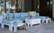 An outdoor patio showcases a pre-assembled base with the Mainstay Sectional Relaxed Middle by Ledge Lounger, featuring blue and yellow cushions. A centered white coffee table holds drinks. Nearby, theres a straw hat, two-toned bag, and sandals. Reflections in the glass wall reveal trees.
