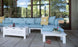A peaceful outdoor patio features a Mainstay Sectional Relaxed Endcap in light blue by Ledge Lounger, enhanced with yellow-patterned pillows. A white sectional table holds drinks and a tray, while sandals suggest leisure. Large windows reveal lush greenery beyond.