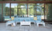 A modern patio features the Mainstay Sectional by Ledge Lounger in light blue with a cushion set of yellow and white pillows. A sleek white coffee table holds drinks, while a striped bag and sun hat are nearby. Glass doors beautifully reflect the lush green landscape.