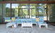 A modern patio features the Mainstay Sectional by Ledge Lounger in light blue with a cushion set of yellow and white pillows. A sleek white coffee table holds drinks, while a striped bag and sun hat are nearby. Glass doors beautifully reflect the lush green landscape.
