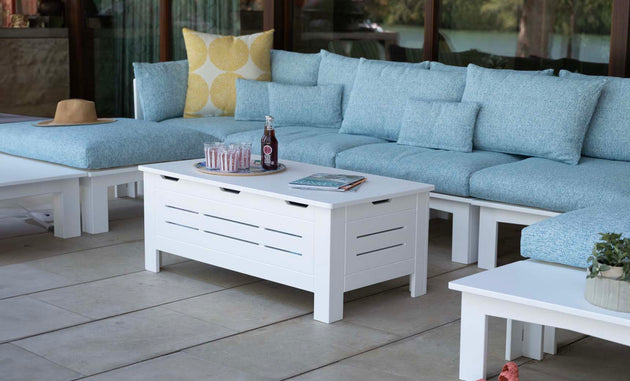 The outdoor patio includes a light blue Mainstay Sectional sofa, a white coffee table with a tray holding glasses, a bottle, and magazines. Nearby is a Mainstay Sectional Relaxed Right Armchair by Ledge Lounger, set beside a beige hat on the bench amid serene greenery and water.