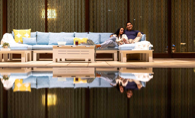 A couple unwinds on a Ledge Lounger Mainstay Sectional Relaxed Middle under warm lighting, enjoying drinks at the table. Their reflection dazzles in the pool, amid a cozy ambiance enhanced by plush cushions that invite serenity and peace.