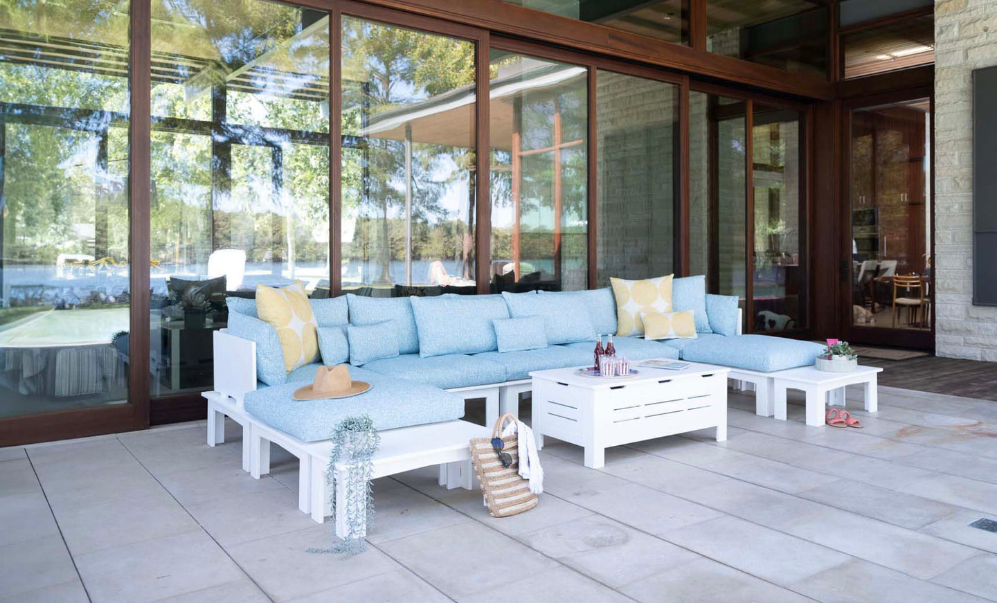 The outdoor patio showcases the Mainstay Sectional Relaxed Left Corner by Ledge Lounger in light blue, adorned with yellow and white cushions. A white coffee table and two side tables perfect the scene, with large glass doors reflecting trees and offering a tranquil lake view.