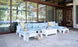 The outdoor patio showcases the Mainstay Sectional Relaxed Left Corner by Ledge Lounger in light blue, adorned with yellow and white cushions. A white coffee table and two side tables perfect the scene, with large glass doors reflecting trees and offering a tranquil lake view.