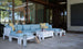 Relax in your outdoor patio with the Mainstay Sectional Relaxed Endcap by Ledge Lounger. The white and blue sectional sofa, adorned with yellow pillows, is complemented by a sectional table holding drinks and a book. A hat, bag, and tinsel add decoration while glass windows reflect trees for tranquility.