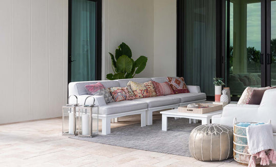 A stylish outdoor space includes a chic Mainstay Sectional 3 Piece Sofa by Ledge Lounger, featuring a long white sofa with colorful patterned cushions. A low white table, decorative lanterns, and a silver pouf accompany it. A large potted plant stands beside the sofa, framed by glass doors.