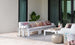 A stylish outdoor space includes a chic Mainstay Sectional 3 Piece Sofa by Ledge Lounger, featuring a long white sofa with colorful patterned cushions. A low white table, decorative lanterns, and a silver pouf accompany it. A large potted plant stands beside the sofa, framed by glass doors.