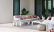 A stylish outdoor space includes a chic Mainstay Sectional 3 Piece Sofa by Ledge Lounger, featuring a long white sofa with colorful patterned cushions. A low white table, decorative lanterns, and a silver pouf accompany it. A large potted plant stands beside the sofa, framed by glass doors.