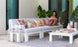 A woman relaxes on the Mainstay Sectional 3 Piece Sofa by Ledge Lounger, adorned with decorative pillows. She sips a drink and holds a tablet on a patio with large glass doors, a rug, and tall candles glowing in lanterns beside her.