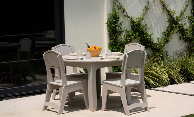 The Mainstay Round Dining Table by Ledge Lounger, complete with an umbrella sleeve, is set on a patio with four beige chairs. It holds plates, glasses, and a bowl of oranges and is placed near a vine-covered wall beside a large window reflecting this serene setting.