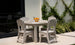 The Mainstay Round Dining Table by Ledge Lounger, complete with an umbrella sleeve, is set on a patio with four beige chairs. It holds plates, glasses, and a bowl of oranges and is placed near a vine-covered wall beside a large window reflecting this serene setting.
