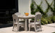The Mainstay Round Dining Table by Ledge Lounger, complete with an umbrella sleeve, is set on a patio with four beige chairs. It holds plates, glasses, and a bowl of oranges and is placed near a vine-covered wall beside a large window reflecting this serene setting.